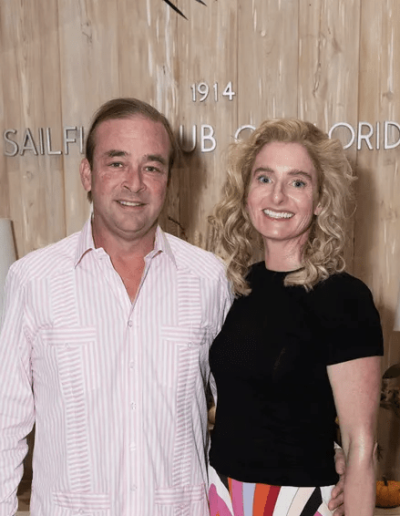 A man and a woman stand together, smiling, in front of a wooden wall with "Sailfish Club of Florida" written on it. Two lamps are visible on either side of them.